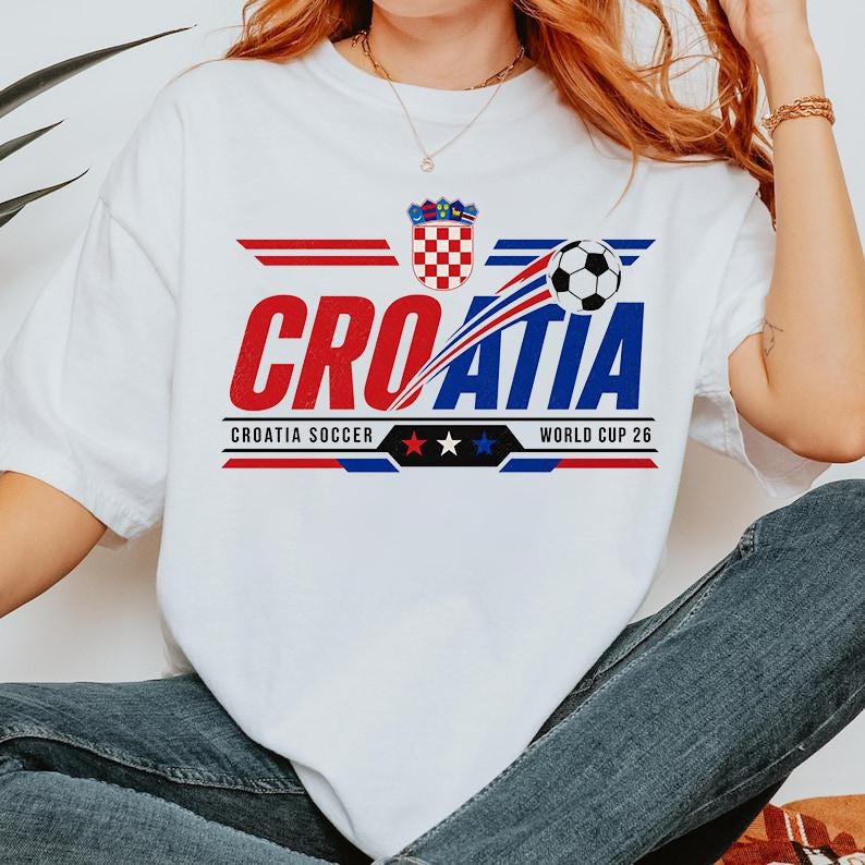 May include: A white short-sleeved t-shirt featuring a graphic design with the text CROATIA, the Croatian coat of arms, a soccer ball, and the text CROATIA SOCCER WORLD CUP 26 with three stars in red, white, and blue colors on the front.