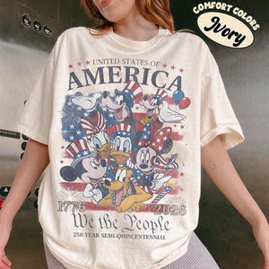 May include: An ivory t-shirt featuring a graphic of cartoon characters wearing patriotic hats against a faded American flag background. Text includes UNITED STATES OF AMERICA, 1776, 2026, We the People, and 250-YEAR SEMI-QUINCENTENNIAL. A sticker reads COMFORT COLORS Ivory.
