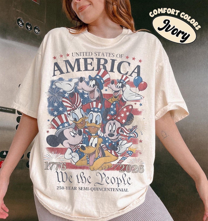 May include: An ivory t-shirt featuring a graphic of cartoon characters wearing patriotic hats against a faded American flag background. Text includes UNITED STATES OF AMERICA, 1776, 2026, We the People, and 250-YEAR SEMI-QUINCENTENNIAL. A sticker reads COMFORT COLORS Ivory.