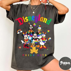 Puede incluir: Camiseta gris oscuro con la palabra "Disneyland" en colores arco&iacute;ris y personajes de dibujos animados como Mickey Mouse y Donald Duck. El texto "Pride Nite" est&aacute; en la parte inferior.