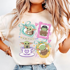 K&ouml;nnte beinhalten: Cremefarbenes T-Shirt mit Grogu, auch bekannt als Baby Yoda, in vier verschiedenen gerahmten Illustrationen. Jeder Rahmen hat ein einzigartiges Design, einschlie&szlig;lich Schleifen, Herzen und einer Teetasse. Das Wort "GROGU" ist in einer verspielten Schrift geschrieben.