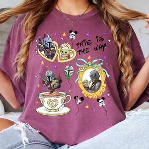 May include: A maroon t-shirt featuring illustrations of characters from a popular space western. The design includes framed portraits, a teacup, and the phrase "This is the Way". Decorative elements include stars and character-themed balloons.