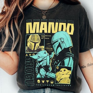 May include: Dark gray t-shirt featuring a graphic design with the text "MANDO" in yellow block letters. The design includes illustrations of a helmeted character and a small green creature, with the text "GROGU" and "THE LEGEND CONTINUES".