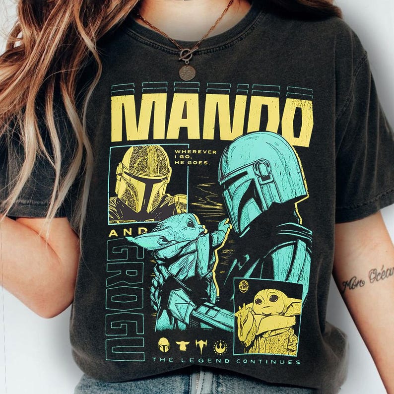 May include: Dark gray t-shirt featuring a graphic design with the text "MANDO" in yellow block letters. The design includes illustrations of a helmeted character and a small green creature, with the text "GROGU" and "THE LEGEND CONTINUES".