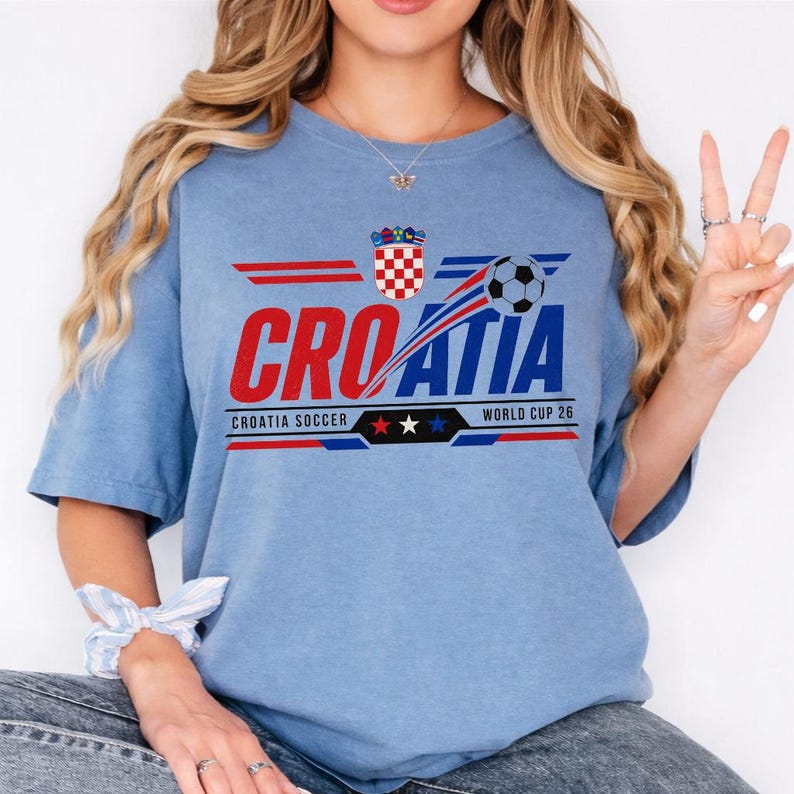 May include: A light blue short-sleeve t-shirt featuring a graphic that reads CROATIA in red and blue letters. The design includes the Croatian coat of arms, a soccer ball, and the text CROATIA SOCCER WORLD CUP 26 with three small stars.