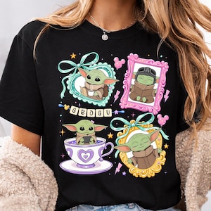 K&ouml;nnte beinhalten: Schwarzes T-Shirt mit vier gerahmten Illustrationen von Grogu, auch bekannt als Baby Yoda. Die Rahmen sind mit Schleifen, Sternen und Mickey Mouse-Silhouetten verziert. Das Wort "GROGU" ist in einer verspielten Schriftart geschrieben.
