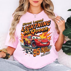 May include: Pink t-shirt featuring a graphic of Lightning McQueen from the movie Cars. The design includes the text "Street Legend Lightning McQueen" and "95 Need for Speed Race Ready". The car is red with the number 95.
