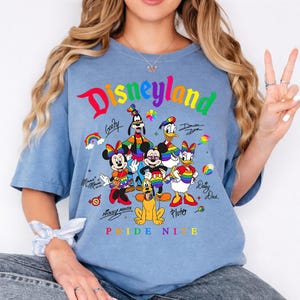 Puede incluir: Una camiseta azul claro con la palabra "Disneyland" en colores arco&iacute;ris, junto con personajes de dibujos animados como Mickey Mouse, Minnie Mouse, Donald Duck y Pluto. La camiseta tambi&eacute;n tiene el texto "Pride Nite".