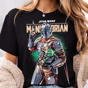 Pu&ograve; includere: T-shirt nero con grafica di Star Wars con The Mandalorian e Grogu. Il design include la scritta "STAR WARS" sopra "MANDALORIAN" in beige. Il Mandaloriano &egrave; in piena armatura, con un blaster.