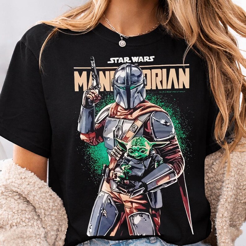 Pu&ograve; includere: T-shirt nero con grafica di Star Wars con The Mandalorian e Grogu. Il design include la scritta "STAR WARS" sopra "MANDALORIAN" in beige. Il Mandaloriano &egrave; in piena armatura, con un blaster.