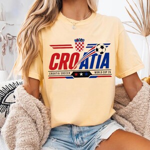 May include: A pale yellow short-sleeve t-shirt featuring a graphic print with the text CROATIA, a soccer ball, the Croatian coat of arms, and the words CROATIA SOCCER WORLD CUP 26 with three stars in red, white, and blue colors on the front.