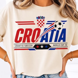 May include: A cream-colored t-shirt featuring a graphic with the text CROATIA in red and blue letters. The design includes the Croatian coat of arms, a soccer ball, and the text CROATIA SOCCER WORLD CUP 26 with red, white, and blue stars.