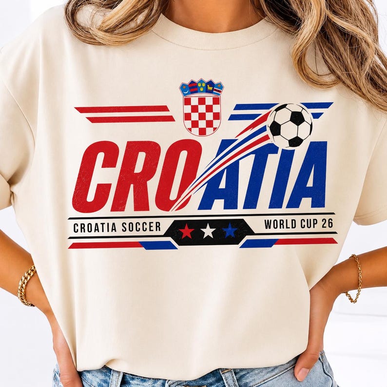 May include: A cream-colored t-shirt featuring a graphic with the text CROATIA in red and blue letters. The design includes the Croatian coat of arms, a soccer ball, and the text CROATIA SOCCER WORLD CUP 26 with red, white, and blue stars.