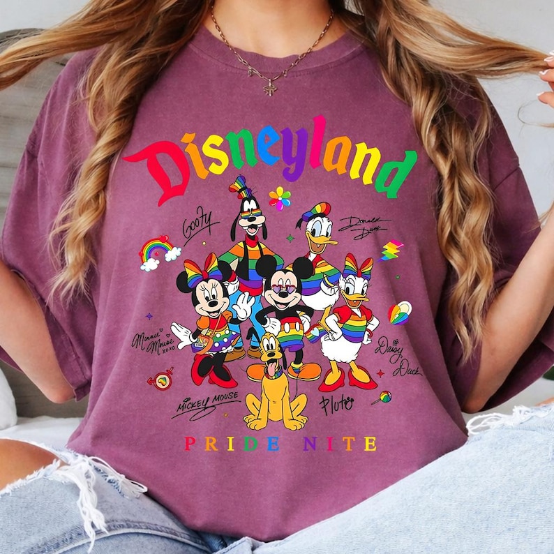Puede incluir: Una camiseta granate con la palabra "Disneyland" en colores arco&iacute;ris. La camiseta presenta personajes de dibujos animados como Mickey Mouse, Minnie Mouse, Donald Duck y Goofy, con el texto "Pride Nite" en colores arco&iacute;ris.