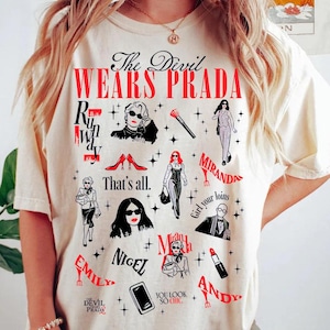 May include: Cream-colored t-shirt featuring the text "The Devil Wears Prada" in black and red, along with various illustrations and character names from the movie. The design includes images of sunglasses, lipstick, and fashion figures.