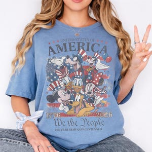 May include: A blue t-shirt featuring a graphic print of cartoon characters wearing patriotic hats. The text reads UNITED STATES OF AMERICA, 1776 2026, We the People, and 250-YEAR SEMI-QUINCENTENNIAL. The design includes fireworks and a flag motif.