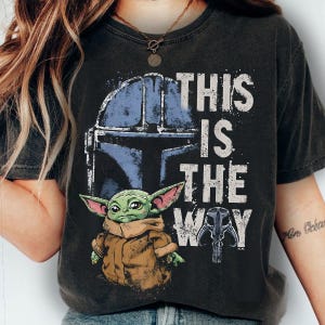 May include: Dark grey t-shirt with a graphic of a blue Mandalorian helmet, a green alien, and the text "THIS IS THE WAY" in white. The shirt has a vintage, worn appearance.