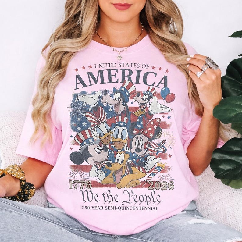 May include: A light pink t-shirt featuring a graphic of cartoon characters wearing patriotic hats. The text reads UNITED STATES OF AMERICA, 1776, 2026, We the People, and 250-YEAR SEMI-QUINCENTENNIAL. The design includes a faded American flag and fireworks.