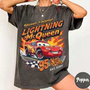 May include: Dark gray t-shirt featuring Lightning McQueen from the movie Cars. The design includes the text "Street Legend Lightning McQueen" and "95 Need for Speed Race Ready" with a cartoon image of the red race car. The shirt has a vintage, worn look.