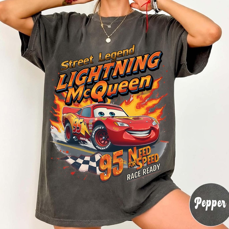 May include: Dark gray t-shirt featuring Lightning McQueen from the movie Cars. The design includes the text "Street Legend Lightning McQueen" and "95 Need for Speed Race Ready" with a cartoon image of the red race car. The shirt has a vintage, worn look.