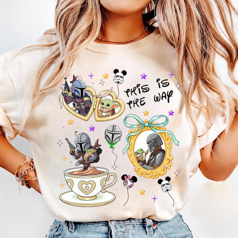 May include: Cream-colored graphic tee featuring illustrations of characters from a popular space western. The design includes the text "This is the Way" and images of a Mandalorian, Grogu, and other themed elements. The tee has a crew neck.