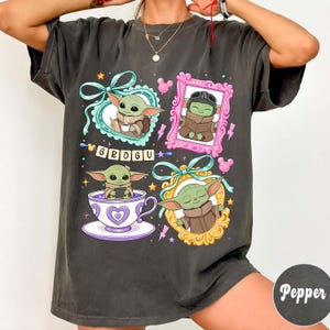 K&ouml;nnte beinhalten: Dunkelgraues T-Shirt mit Grogu aus The Mandalorian. Das Shirt hat vier gerahmte Illustrationen von Grogu, mit dem Wort "GROGU" in der Mitte. Die Illustrationen sind in t&uuml;rkisen, rosa und goldenen Rahmen.