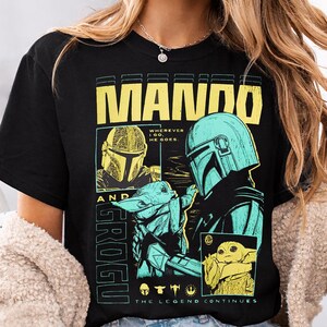 May include: Black t-shirt featuring a graphic design with the text "MANDO AND GROGU". The design includes images of a helmeted character and a small green creature. The text "THE LEGEND CONTINUES" is also present.