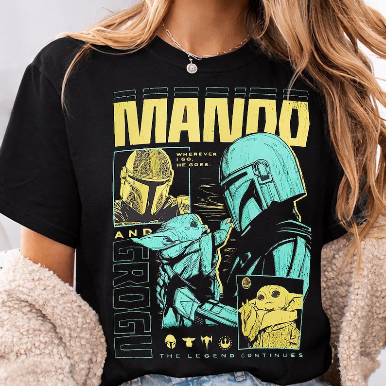 May include: Black t-shirt featuring a graphic design with the text "MANDO AND GROGU". The design includes images of a helmeted character and a small green creature. The text "THE LEGEND CONTINUES" is also present.