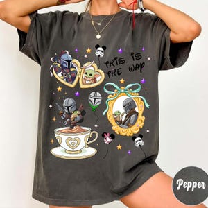 May include: Dark gray t-shirt featuring cartoon illustrations of characters from a popular space western. The design includes images within heart-shaped frames, a teacup, and balloons. The text "THIS IS THE WAY" is also present.