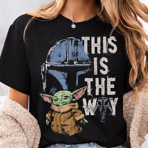 May include: Black t-shirt with a graphic of a Mandalorian helmet, a green alien, and the phrase "THIS IS THE WAY" in white distressed lettering. The design includes blue and brown tones.