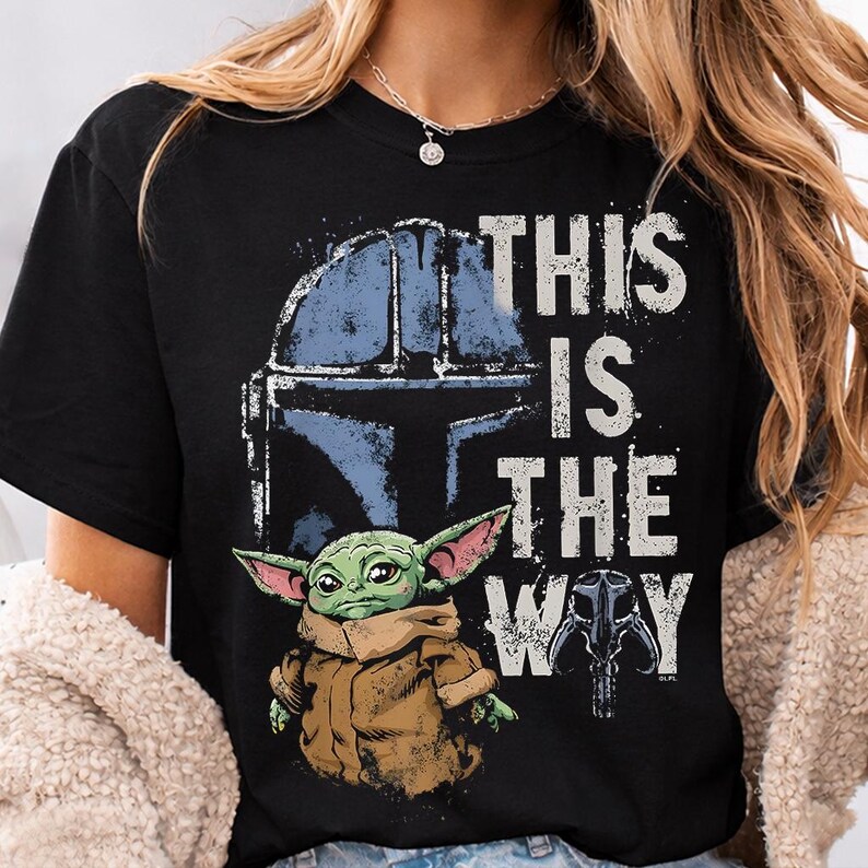 May include: Black t-shirt with a graphic of a Mandalorian helmet, a green alien, and the phrase "THIS IS THE WAY" in white distressed lettering. The design includes blue and brown tones.