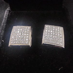 May include: A pair of square silver earrings, each encrusted with numerous small, clear gemstones. The earrings are set against a black velvet background, suggesting a jewelry display or storage case. The gemstones sparkle, catching the light.