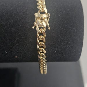 May include: A gold-colored curb chain bracelet with a secure clasp. The bracelet is displayed on a black surface, showcasing its polished links and intricate closure mechanism. The chain links are thick and closely interlocked.