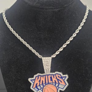 May include: A silver-toned rope chain necklace featuring a pendant with the New York Knicks logo. The pendant is outlined in clear rhinestones and displays the word KNICKS in blue and orange, with a basketball graphic at the bottom center.