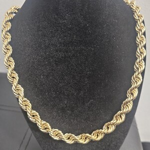 May include: A gold-colored rope chain necklace displayed on a black velvet bust. The chain is made of tightly twisted links, creating a thick, textured appearance. The necklace is a classic jewelry piece.