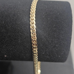 May include: A gold-colored curb chain bracelet. The bracelet is displayed against a black background. The chain links are closely connected, creating a smooth, continuous surface. The bracelet is a classic jewelry piece.