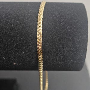 May include: A gold-colored chain bracelet with a flat curb link design. The bracelet is displayed against a black background, showcasing its shiny, reflective surface. The chain is approximately 7 inches long.