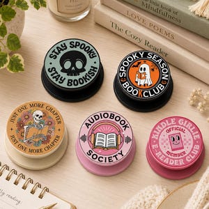May include: Five circular phone grips with various designs: a light green one with a skull and text 'STAY SPOOKY STAY BOOKISH', an orange one with a ghost reading and 'SPOOKY SEASON BOOK CLUB', a tan one with a skeleton reading, a pink one with headphones and an open book, and a pink one with a smiling e-reader.