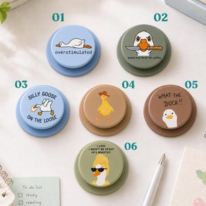 May include: Six circular phone grips featuring various goose illustrations and text. Designs include a goose with a knife, a goose in a towel, and a goose wearing a hat. The colors are light blue, olive green, tan, and brown, each labeled with numbers 01 through 06.