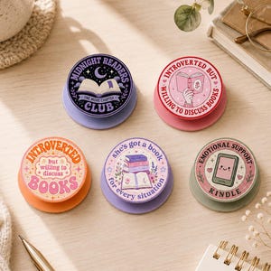 May include: Five circular phone grips featuring book-themed designs. One is purple with a moon and open book, one pink with a person reading, one orange with retro text, one lavender with a stack of books, and one green with a smiling e-reader.