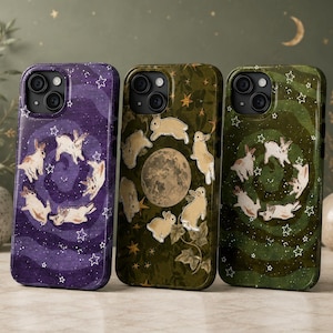 May include: Three smartphone cases featuring rabbit illustrations. The left case is purple with white stars and rabbits. The center case is olive green with a central moon, stars, and rabbits. The right case is dark green with white stars and rabbits.