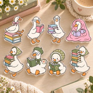 May include: A set of eight acrylic charms featuring white geese in various activities. The geese are depicted reading books, holding stacks of colorful books, wearing bonnets and glasses, and wrapped in a pink blanket while holding a mug.