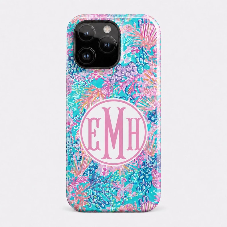 May include: A smartphone case featuring a vibrant, multi-colored pattern of coral, seashells, and aquatic flora in shades of turquoise, pink, and purple. Centered on the case is a white circular emblem containing the pink monogram letters EMH.