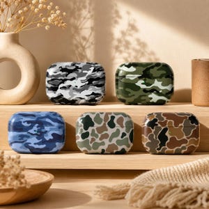 May include: Five rectangular wireless earbud charging cases featuring various camouflage patterns. The cases display distinct color palettes including black and white, forest green, shades of blue, muted earth tones, and brown and tan combinations.