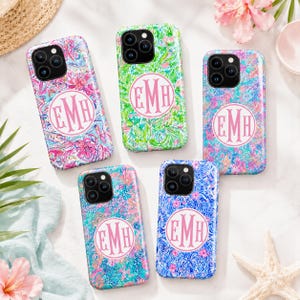 May include: Five smartphone cases featuring vibrant, multi-colored floral and abstract patterns. Each case displays a circular white emblem in the center containing the pink monogram letters EMH. The cases have black camera lens cutouts.