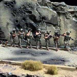 Op de afbeelding: Miniatuur diorama met zeven soldaten in camouflage-uniformen, elk met een geweer, voor een verweerde betonnen muur. Een kleine hond is ook aanwezig. De scène speelt zich af in een woestijnachtige omgeving.