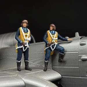 May include: Two miniature pilot figurines standing on a gray aircraft model. Each figure wears a dark blue uniform, a yellow life vest with white straps, brown leather flight helmet with goggles, and black boots. The figures are painted in detail.