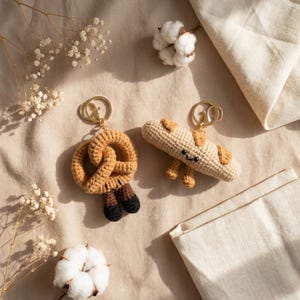 May include: Two handmade crochet keyrings: a pretzel with brown and black details and a baguette with a smiling face and small legs. Both keyrings have gold-coloured hardware. The background is a neutral beige.