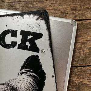 May include: Two metal signs, one partially covering the other. The top sign is white with black text that reads "CK" and a rooster illustration. The sign has a distressed, vintage look with a black border.