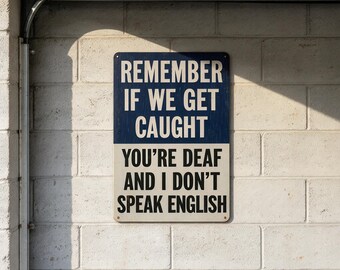 May include: A rectangular metal sign with a two-tone design. The top half is dark blue with white text reading REMEMBER IF WE GET CAUGHT. The bottom half is off-white with black text reading YOU'RE DEAF AND I DON'T SPEAK ENGLISH.